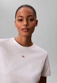 Jeune femme à la peau lisse et aux cheveux attachés porte un T-shirt blanc avec un petit patch logo Calvin Klein rouge et noir.