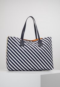 Borsa a tracolla a righe diagonali blu navy e bianca con doppi manici neri e interno arancione, esposta su un piedistallo bianco contro uno sfondo grigio.