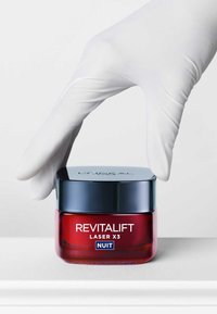 Bocal en verre rouge de Revitalift Laser X3 Nuit avec un couvercle bleu marine, tenu par une main gantée, avec un texte blanc sur l'étiquette avant.