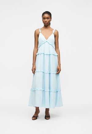 Femme portant une robe longue bleu clair sans manches avec des volants superposés, debout avec une expression neutre et des talons ouverts noirs.
