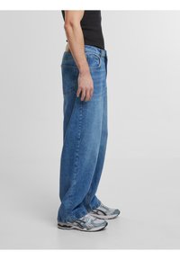 Jean en denim bleu ample avec un délavage, cinq poches et une coupe droite, présentés de côté avec un débardeur noir et des baskets.