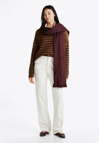 Brązowy sweter w paski granatowe, białe szerokie spodnie oraz bordowa, wzorzysta chusta z frędzlami. Stylizacja zawiera cienki pasek i ciemne buty.