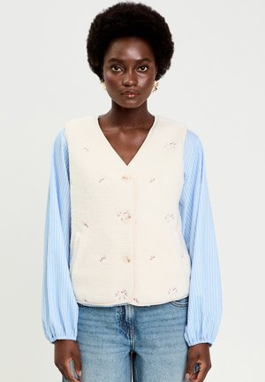 Donna con capelli corti e ricci che indossa un gilet crema ricamato sopra una camicetta a maniche lunghe a righe azzurre e jeans blu, rivolta in avanti.