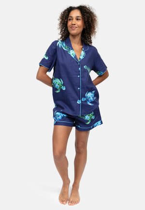 Donna con capelli ricci che indossa una camicia blu navy a maniche corte con bottoni e pantaloncini con stampa di tartarughe marine verdi e blu, in piedi scalza.