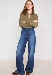 Blouse brodée vert olive avec un décolleté en V, associée à un jean en denim bleu à taille haute et jambes larges, et des mocassins marron.