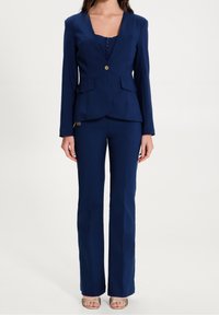 Žena v námořnickém modrém přiléhavém blejzru a odpovídajících širokých kalhotách, doplněná o zlaté sandály s otevřenou špičkou, stojící na bílém pozadí.