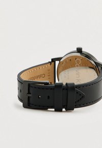 Schwarzes Lederarmband mit kontrastierenden Nähten, Metallverschluss und einem runden Gehäuse, das eine tanfarbene Lederunterlage und das Markenlogo innen zeigt.