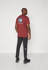 Kestenjasto kratki rukav T-shirt s grafičkim logotipom na leđima i bijelim tekstom. Crne hlače i crna obuća s teksturiranom završnom obradom.