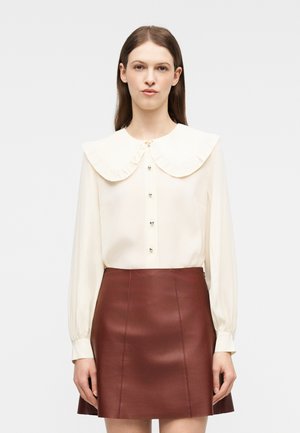 Γυναίκα που φοράει κρέμ μπλούζα με μεγάλο στρογγυλό γιακά και κουμπιά, συνδυασμένη με καφέ δερμάτινη μίνι φούστα σε γραμμή Α, στέκεται μπροστά σε απλό φόντο.