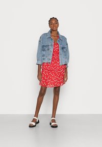 Casaco de ganga sobre um vestido floral vermelho com design em camadas. Sandálias brancas com solas pretas completam o visual. Fundo branco liso.