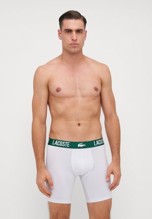 Boxer bianchi con una cintura verde con il logo Lacoste. Il tessuto sembra liscio ed elasticizzato, aderente al corpo. Il modello è in posizione eretta.