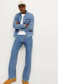 Homem vestindo uma jaqueta de denim azul clara e jeans, camiseta branca, botas castanhas e um boné bege, de pé com os braços cruzados contra um fundo branco.