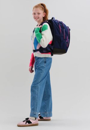 Lächendes Mädchen mit roten Haaren, das einen bunten Pullover, blaue Jeans und rosa Turnschuhe trägt, dazu einen großen dunkelblauen Rucksack vor schlichten Hintergrund.