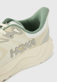 Helle Laufschuhe aus strukturiertem Material mit grünen Akzenten und einem auffälligen grauen Logo. Verfügt über eine gepolsterte Sohle und eine Schnürgestaltung.
