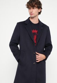 Marineblauer Wollmischmantel mit Reverskragen, zwei Fronttaschen und fünf Knöpfen. Wird über einem rot gemusterten Pullover getragen. Glatte Textur.