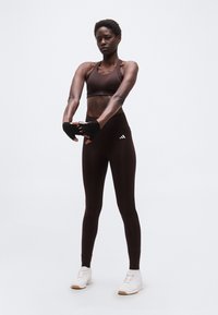 Αθλητική γυναίκα τεντώνει τα χέρια μπροστά, φορώντας μαύρα αθλητικά ρούχα, γάντια χωρίς δάχτυλα και λευκά αθλητικά παπούτσια, στέκεται μπροστά σε απλό φόντο.