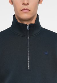Sweatshirt noir zippé avec col montant, tissu texturé et petit logo sur la poitrine gauche. La fermeture éclair est de couleur argentée avec un tirant bleu.