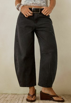 Persoon in wijde zwarte jeans, een zwarte riem, een zilveren armband en bruine platform sandalen op een betegelde vloer.