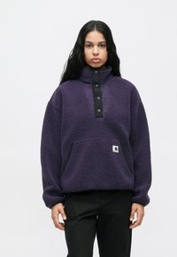 Pikend tumed juuksed omav inimene kannab lillat fliispulloverit mustade nööbiplätudega ja musti pükse, seistes tasase valge tausta ees.