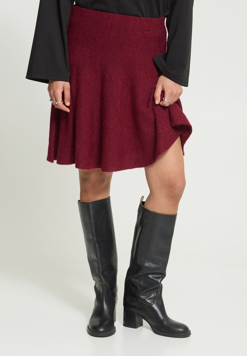 Personne portant une jupe bordeaux évasée, un haut noir à manches longues et des bottes en cuir noir jusqu’aux genoux, debout devant un fond clair.