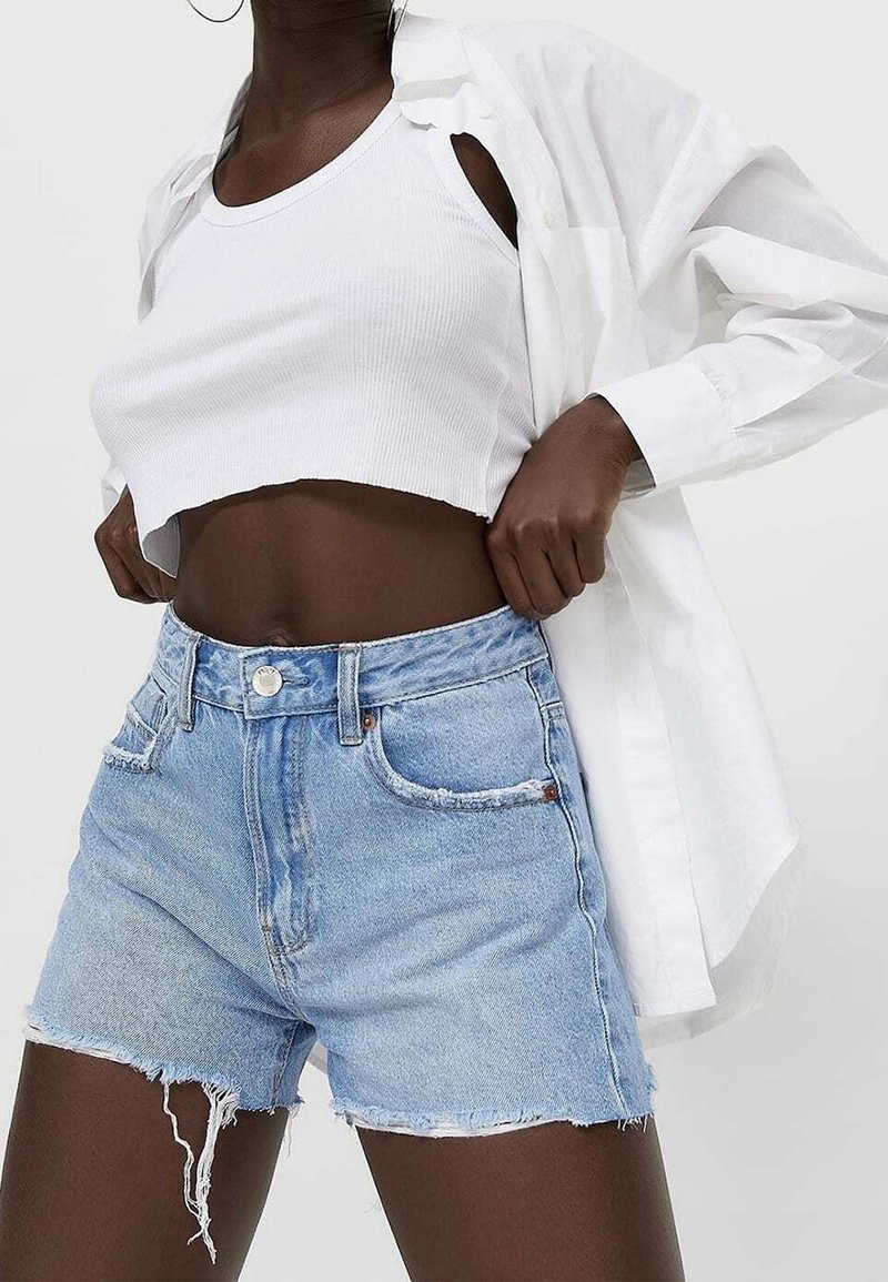 Personne à la peau foncée portant un débardeur court blanc, une chemise blanche ouverte et un short en jean bleu clair effiloché, ajustant les manches de la chemise.