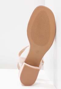 Sandale avec semelle beige et talon bloc, semelle montrant un motif ondulé et marquage taille 37, appuyée contre un mur blanc.