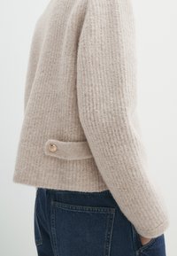 Beige gebreide trui met een gestructureerd patroon, voorzien van een zijsluiting met knopen en een relaxte pasvorm. Gecombineerd met donkerblauwe jeans.