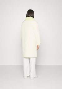 Mujer con chaqueta larga acolchada blanca y cuello verde claro, pantalones anchos blancos y zapatos blancos, de pie de espaldas sobre un fondo liso.