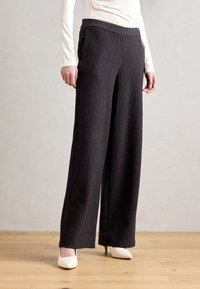 Zwarte, wijde broek met verticale strepen, elastische tailleband en zachte stof. Gecombineerd met witte hoge hakken, geplaatst op een houten vloer.
