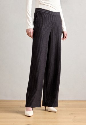 Black, wide-leg pants with vertical pinstripes, elastic waistband, and soft fabric. Paired with white heeled shoes, positioned on wooden floor.