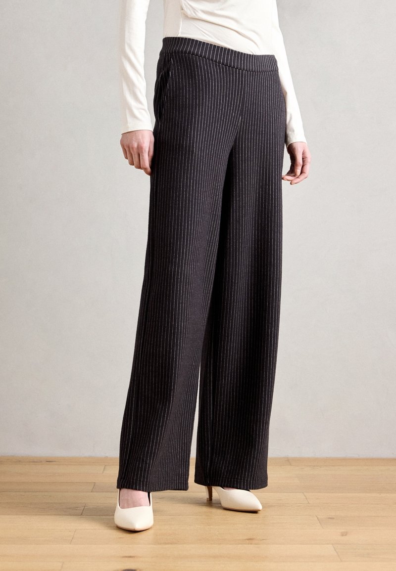 Zwarte, wijde broek met verticale strepen, elastische tailleband en zachte stof. Gecombineerd met witte hoge hakken, geplaatst op een houten vloer.