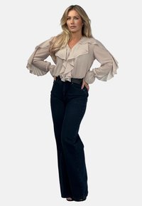 Jasno-beżowa bluzka z falbanami, długimi rękawami i dekoltem w kształcie litery V. Połączona z ciemnoniebieskimi spodniami typu dzwony o wysokim stanie oraz czarnymi sandałami.