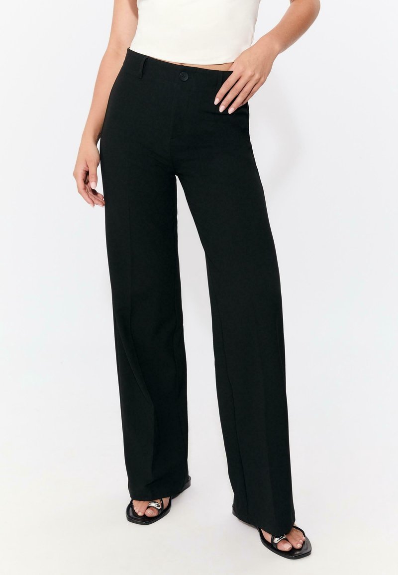 Femme portant un pantalon noir taille haute à jambes larges avec une fermeture à bouton et zip, associé à des sandales noires ouvertes et un haut sans manches blanc.