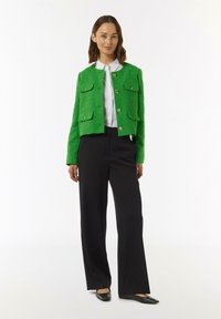 Groene tweed cropped jack met twee grote voorzakken, gouden knopen en een gestructureerde afwerking, gecombineerd met zwarte broek met wijde pijpen en platte schoenen.