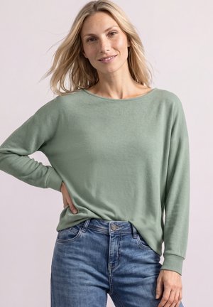 Kvinde med blondt hår iført en lysegrøn langærmet top og blå jeans, smilende med hånden på hoften.