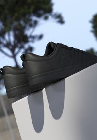 Paire de baskets noires basses avec semelles texturées reposant sur une surface blanche en extérieur avec des arbres flous en arrière-plan.