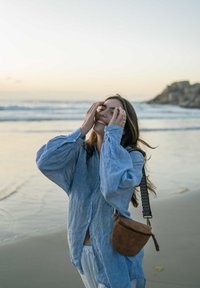 Junge Frau in einem blauen Shirt, lächelt mit geschlossenen Augen und berührt ihr Gesicht an einem sandigen Strand in der Nähe sanfter Meereswellen bei Sonnenuntergang.