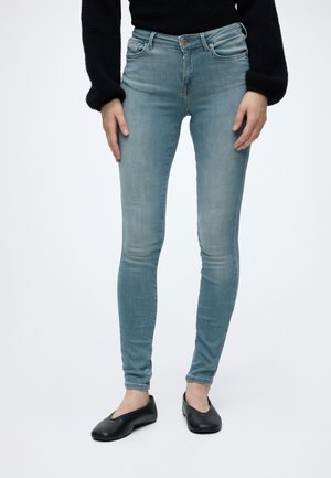 Person iført lyseblå skinny jeans, sort langærmet top og sorte balletsko, der står mod en ensfarvet baggrund.