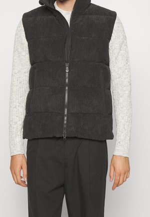 Homme portant une doudoune sans manches en velours côtelé noir avec fermeture éclair frontale sur un pull en maille torsadée gris clair et un pantalon noir.