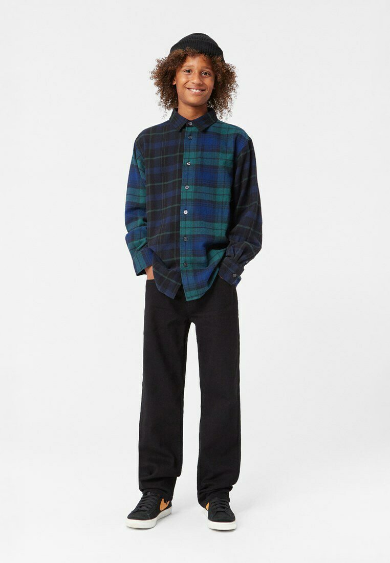 Camicia di flanella a quadri verde e blu navy con colletto a bottoni, abbinata a pantaloni neri e sneakers nere con accenti arancioni.