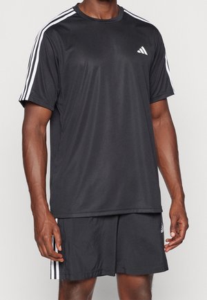 Homme portant un t-shirt de sport noir à manches courtes et un short avec des bandes blanches aux épaules et sur les côtés sur fond uni.