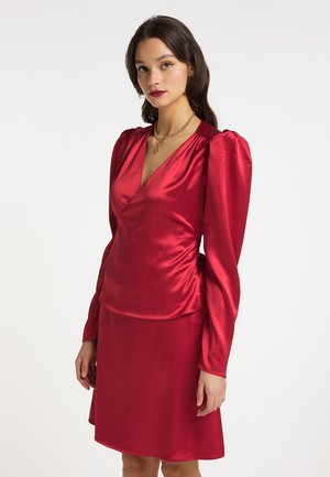 Frau trägt ein glänzendes rotes Langarmkleid mit Puffärmeln und V-Ausschnitt, accessoriert mit einer goldenen Halskette.