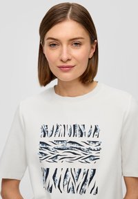 Kobieta z prostymi brązowymi włosami nosi białą koszulkę z czarnym i srebrnym wzorem w paski zebry z cekinami z przodu.