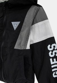 Giacca a vento nera e grigia con cappuccio, chiusura con zip, strisce bianche e una toppa con logo in grandi lettere sulla manica.