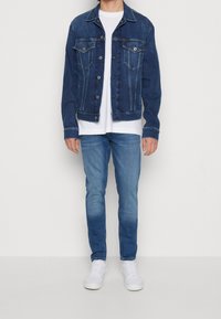 Jeansjacka i mörkblått, med två bröstfickor, knappstängning och klassisk krage. Bärs över en vit t-shirt och ljusblå jeans.