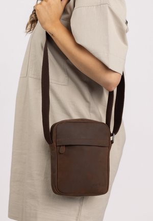 Brown leather crossbody bag with a zippered top, flat front pocket, and a dark fabric strap, shown against a neutral background.