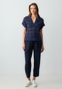 Navyblaues Kurzarmhemd mit V-Ausschnitt, das ein strukturiertes Streifenmuster aufweist. Kombiniert mit lockeren navyblauen Hosen und weißen Sneakers.
