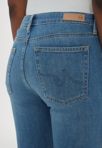 Jean en denim bleu à taille haute, avec un patch en cuir à l'arrière de la ceinture et deux poches arrière avec des coutures décoratives.