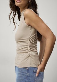 Beige ærmeløs top med en tætsiddende design, samlet tekstur på siden og en rund halsudskæring, parret med blå denim jeans.