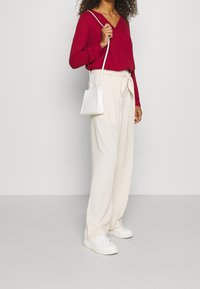 Blusa rossa a maniche lunghe con scollatura a V, abbinata a pantaloni larghi a vita alta color crema e sneakers bianche. Piccola borsa a tracolla bianca.